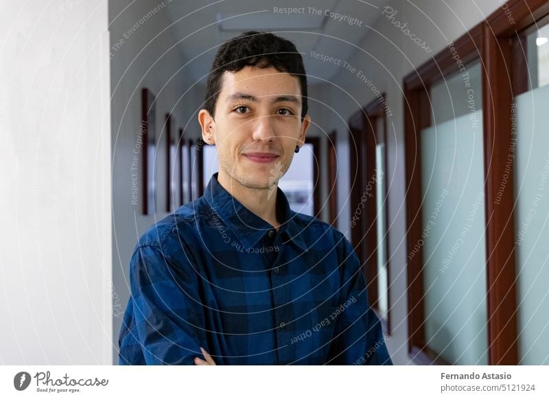 Portrait of a smiling young Caucasian man with his hands crossed in a long hallway wearing a blue and black checked shirt. Portrait photography. Model. Boy with earrings.