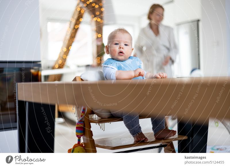 Happy infant sitting at dining table and playing with his toy in traditional scandinavian designer wooden high chair in modern bright atic home superwised by his mother.