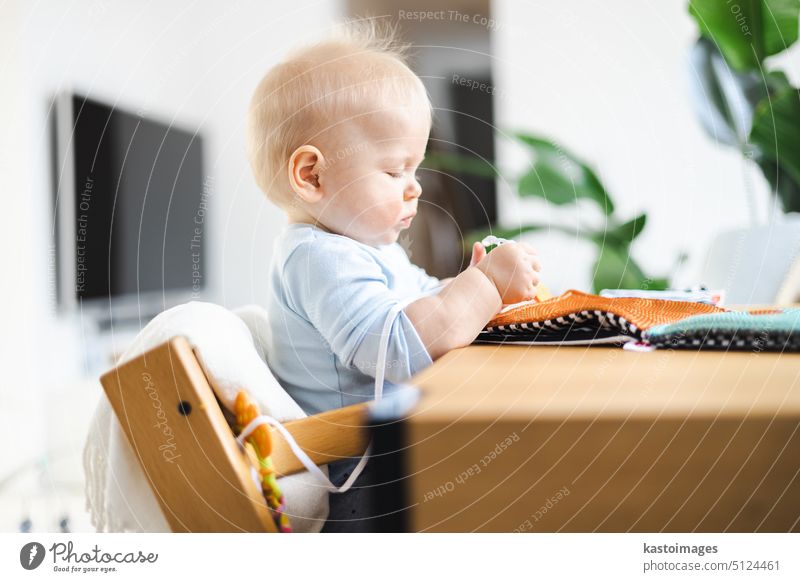 Happy infant sitting at dining table and playing with his toy in traditional scandinavian designer wooden high chair in modern bright atic home. Cute baby playing with toys