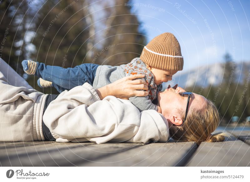 Happy family. Young mother playing with her baby boy infant oudoors on sunny autumn day. Portrait of mom and little son on wooden platform by lake. Positive human emotions, feelings, joy.