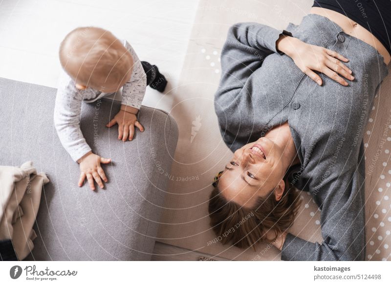 Happy family moments. Mother lying comfortably on children's mat playing with her baby boy watching and suppervising his first steps. Positive human emotions, feelings, joy.