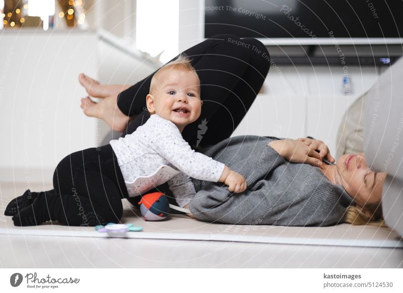 Happy family moments. Mother lying comfortably on children's mat playing with her baby boy watching and suppervising his first steps. Positive human emotions, feelings, joy.