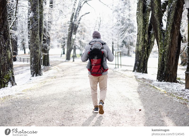 Rear view of sporty father carrying his infant son wearing winter jumpsuit and cap in backpack carrier walking in city park in winter hiking travel parent