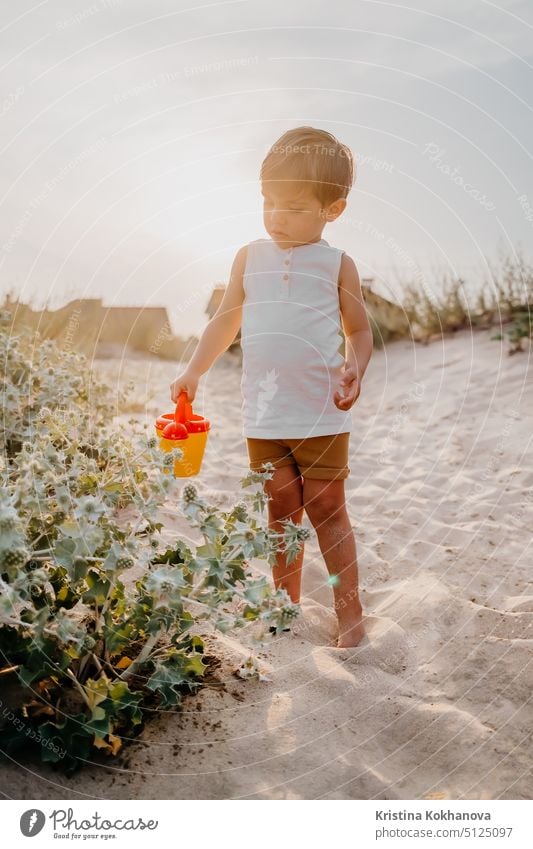 Cute little boy watering plants with can on sandy beach. summer sunny day. Toddler with colorful pot. Natural aestetic portrait of child. fun person vacation