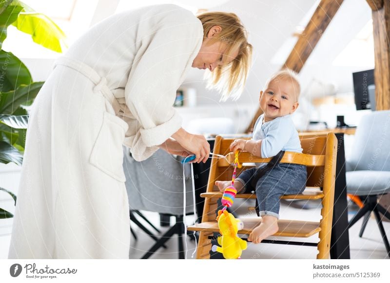 Happy infant sitting and playing with his toy in traditional scandinavian designer wooden high chair in modern bright atic home suppervised by his mother baby