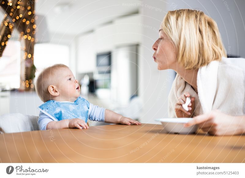 Mother motivating her baby boy infant child while spoon feeding him sitting in high chair at dinning table at home. Baby solid food introduction concept eating