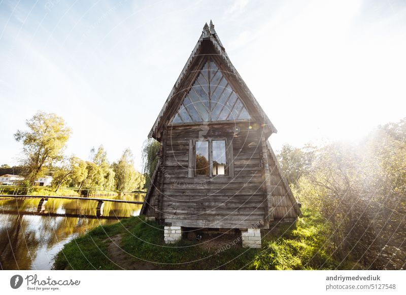 An old wooden village house stands on the edge of the forest by the lake against the blue sky. Old village hut. Peasant dwelling. Abandoned housing people abandoned the house.