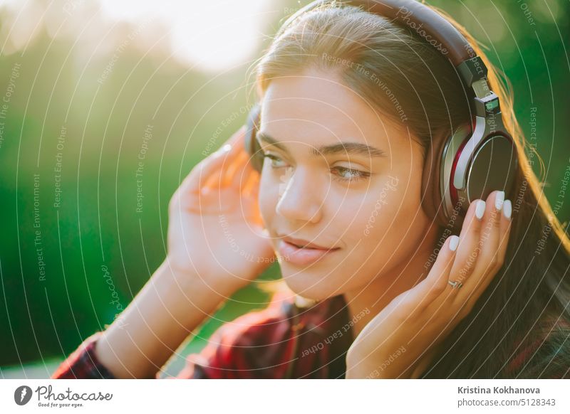 Cute girl's face with headphones. Young woman enjoys music while sitting in green park. Concept of youth, freedom, technology person earphones outdoor listening