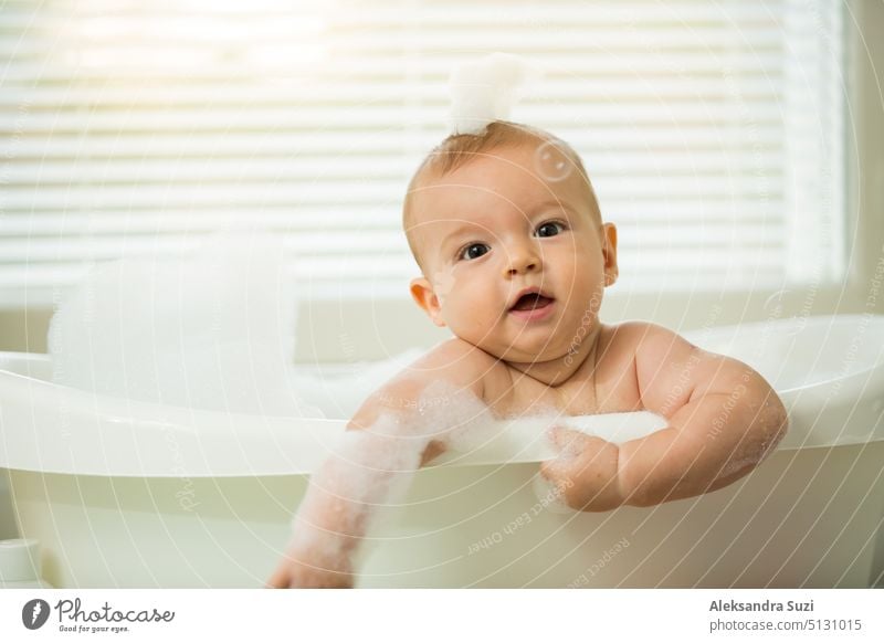 Cute little baby sitting in white bathtub with foam and soap bubbles. Taking bath and playing with toys. Baby hygiene. 6 month 6 month baby adorable bathe