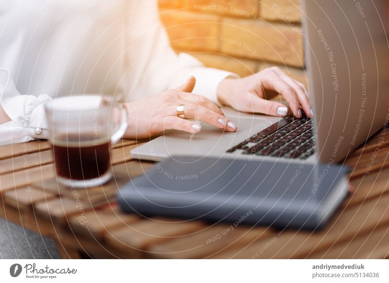 Freelancer girl working at the cafe. Cropped photo of woman hands are using laptop while sitting outdoor. On table glass cup of hot coffee and paper notebook. Coffee break. Online job or studying