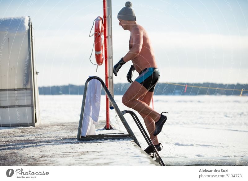 A man in warm hat and gloves swimming in an ice hole, coming out of the water. Winter activities in Finland. Healthy lifestyle active activity adult alternative