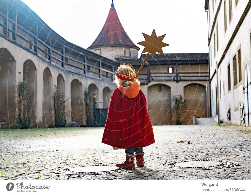A small child from behind with red cape and crown on a large castle square is walking towards a tower carrying a golden star on a stem in front of him