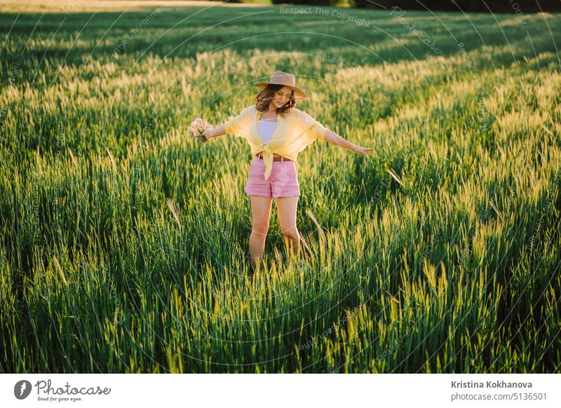 Happy woman in straw hat dancing in fresh green wheat field. Grass background. Amazing nature, farmland, growing cereal plants. Stylish lady with eyewear.