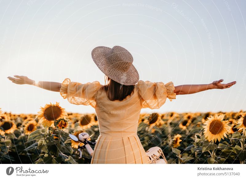 Unrecognizable woman with open arms in sunflowers field. Yellow colors, warm toning. Free girl in straw hat and retro dress. Vintage timeless fashion, amazing adventure, countryside, rural scene