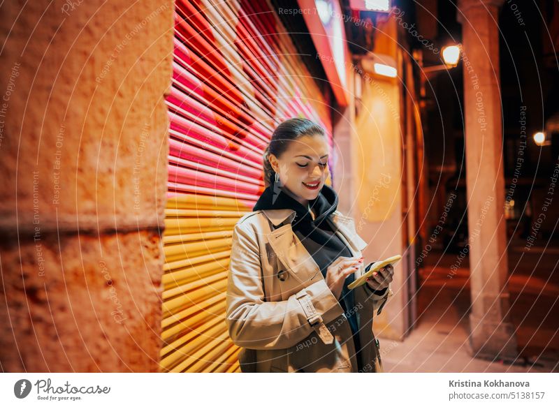 Young woman with smartphone on night city street. Lady standing alone on colorful background, she surfing internet with mobile device. Autumn season. Technology, social apps, connection concept