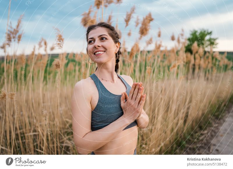 Young woman doing namaste, pranayama yoga breathing exercise on reed natural background. Healthy girl training at summer outdoors. Gratitude, unity with nature, wellness concept.