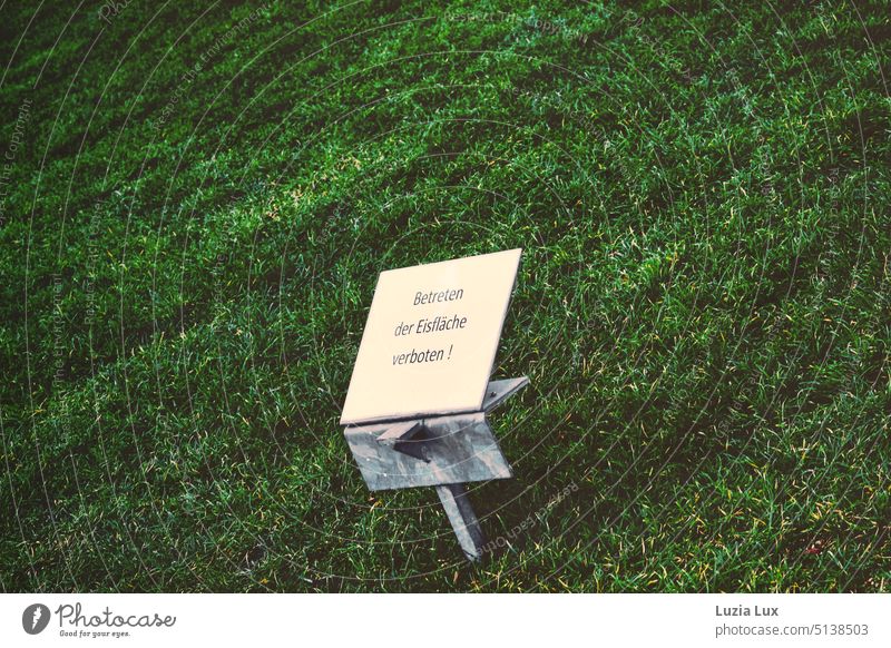 Symbol of climate change: sign 'Do not enter the ice surface!' stands on a green meadow Signs and labeling Frozen surface Winter Winter mood Green Meadow Lawn