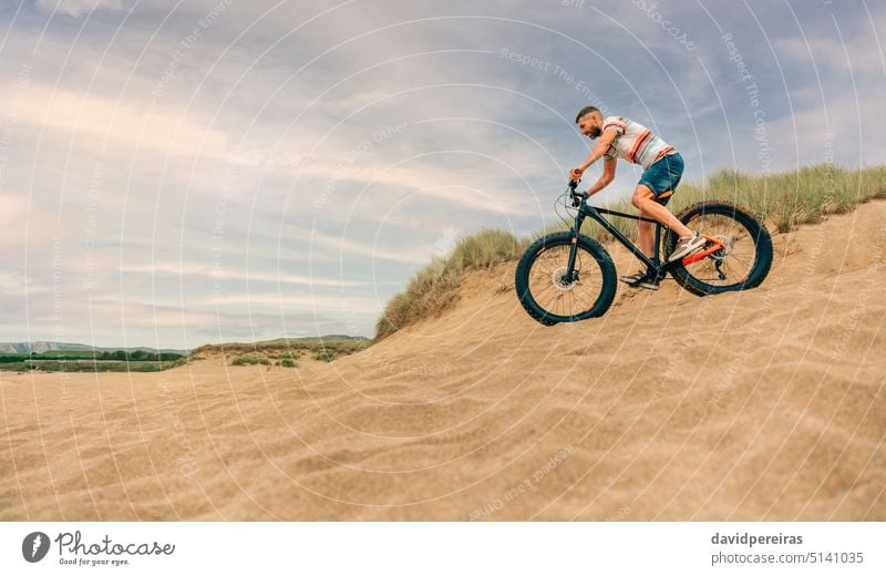 Man riding a fat bike through the beach dunes young man copy space going down person bicycle lifestyle active bicyclist biking profile male guy beard one people