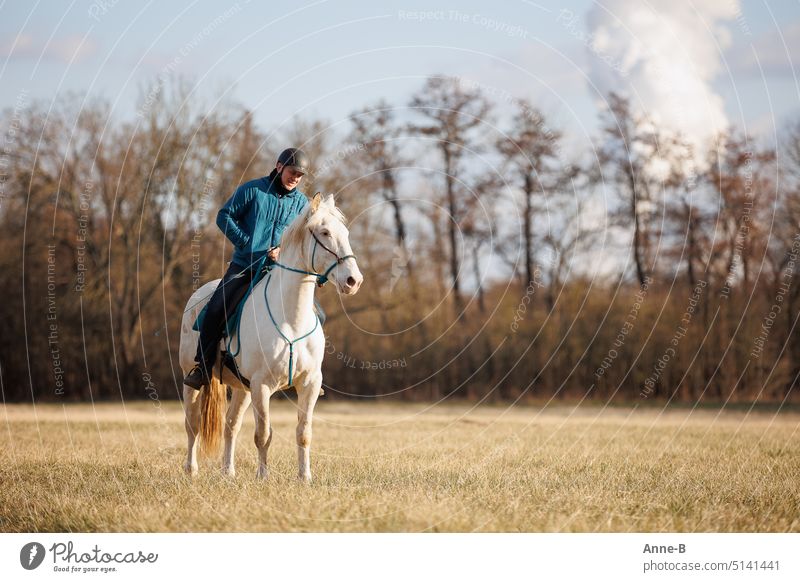 Man sits on a white horse bridled with a bosal and looks for a carrot in his jacket pocket in thanks for the horse carrying him. In the background are blurred autumn trees.