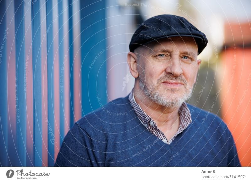 Portrait of a friendly looking man with blue eyes, blue sweater and blue slider cap , beard and direct eye contact. The background is a blue wooden wall.