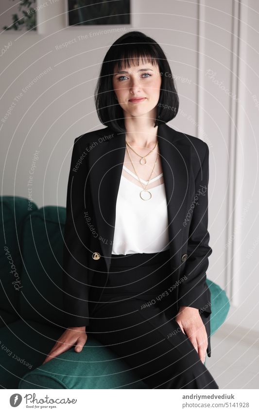 Beautiful business woman in black suit and white blouse is sitting on the green sofa at office, charming brunette female with short hair is looking in camera, girl is working indoors