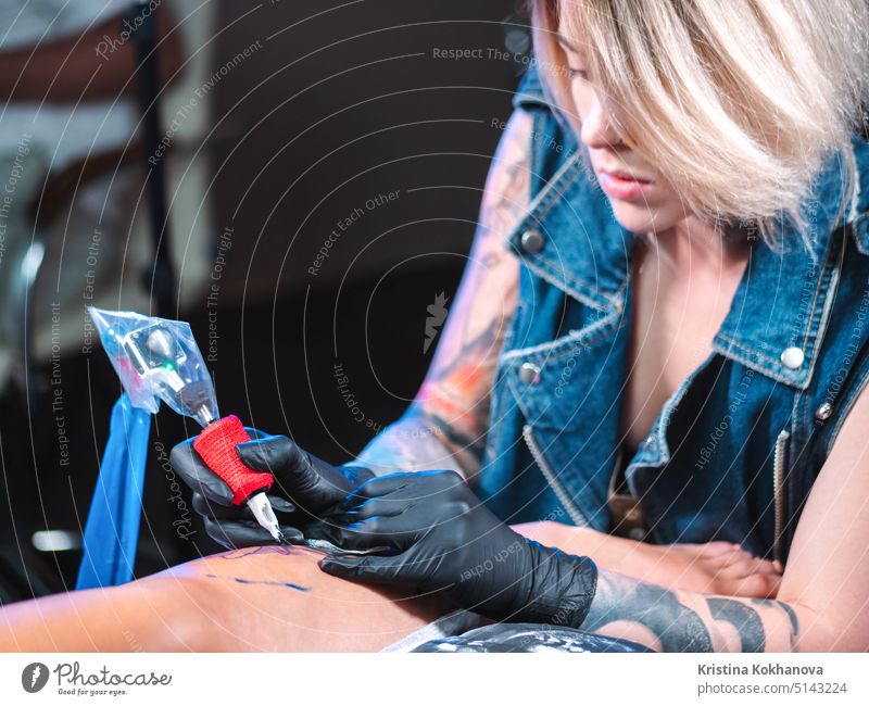 View of professional blonde tattoo female master is painting with black ink, making cover. Works in sterile latex gloves with handmade rotor gun machine in studio.