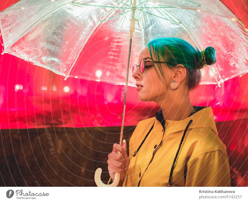 Young pretty girl with blue dyed hair in yellow raincoat and with transparent umbrella stands near fountain. Night neon illumination of city. Portrait of stylish hipster with glasses.
