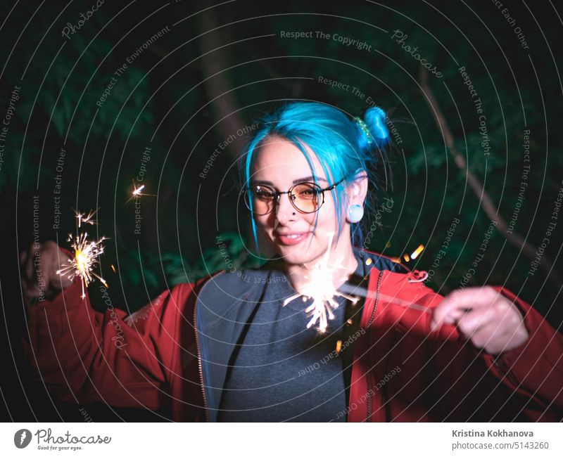 Portrait of hipster girl with blue dyed hair with sparklers in hands and reflection of it in glasses. Close up beautiful young teenage standing at night city, having fun with bengal fire.