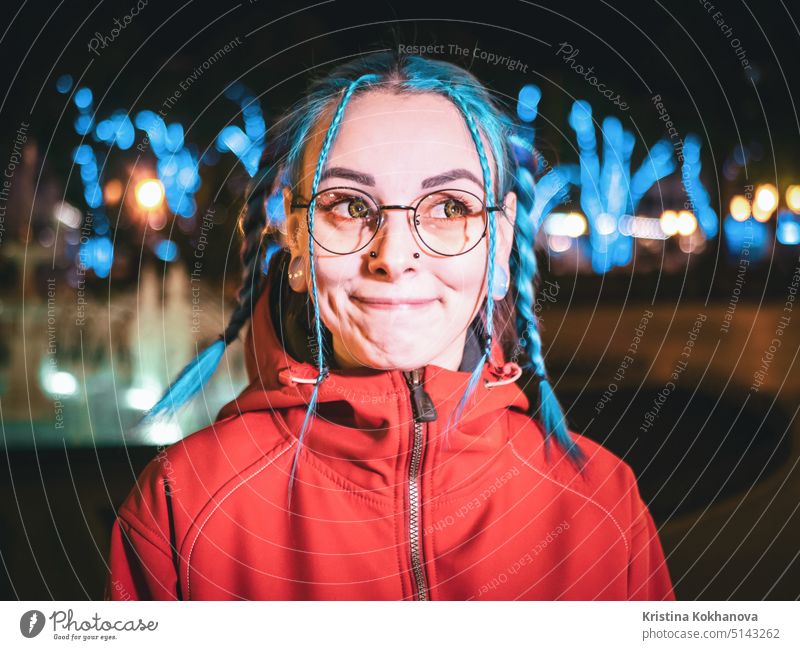 Young beautiful girl with blue dyed unusual hair, transparent glasses, piercing standing at night street with neon lights background. Portrait of happy cute stylish teenager.