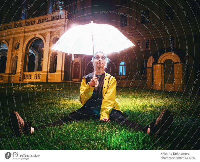 Young pretty girl with blue dyed hair in yellow raincoat and with transparent luminous umbrella sitting on green grass near historic building. Portrait of stylish hipster with glasses.