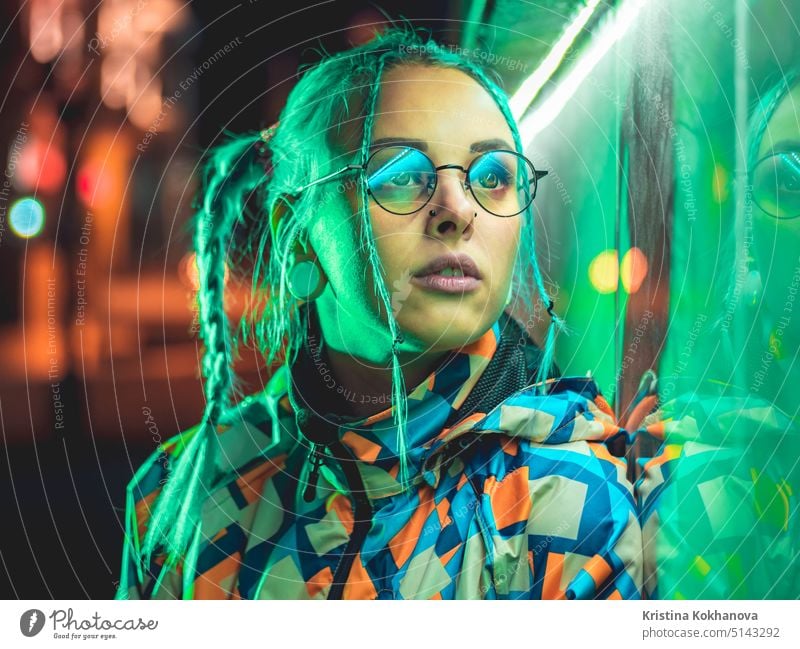 Young pretty girl with unusual hairstyle near glowing green neon light of the city at night. Dyed blue hair in braids. Pensive hipster teenager in glasses.