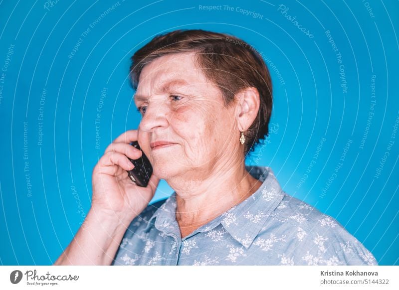Elderly pensioner woman talking with mobile device on blue studio wall. Grandmother smiling. Technology, old people concept. mobile phone grandmother 70s age