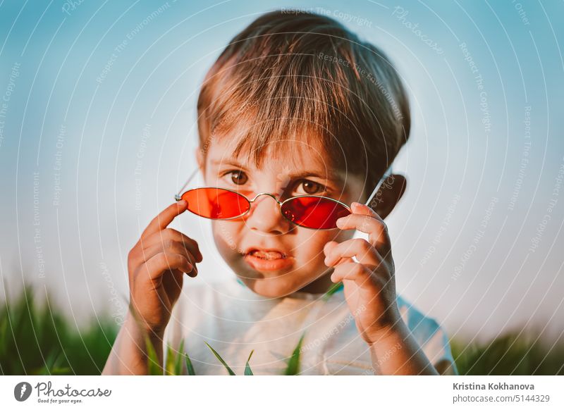 Portrait of funny baby boy in red sunglasses standing in fresh green wheat field. Lovely toddler child explores plants, nature in spring. Childhood, children fashion style concept