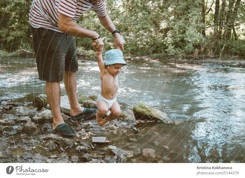 Dad holds his little baby over the river. Funny son tied his legs and does not want to swim in water. family father people child man nature dad kid childhood