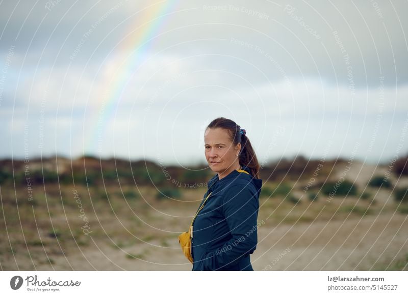 Middle-aged woman looking thoughtful at the camera with a rainbow in the background, concept for future outlook expression standing portrait face copy space