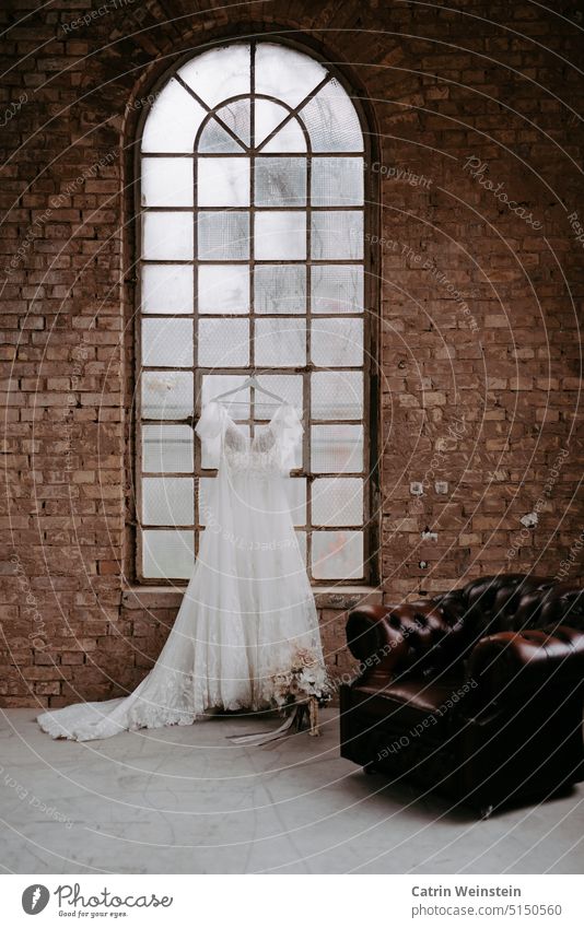 A wedding dress hangs on a hanger by a round-arched window. In front of it is a bridal bouquet and an old leather chair. Brick wall Wedding dress hangers White