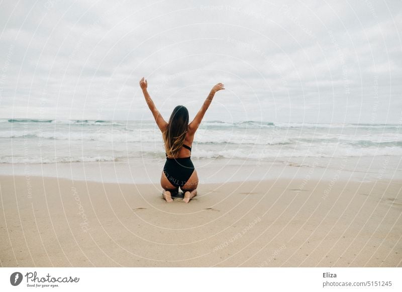 Woman in black swimsuit kneels on beach with arms outstretched and back to camera looking at sea Beach Ocean Swimsuit outstretched arms Joy Freedom vacation