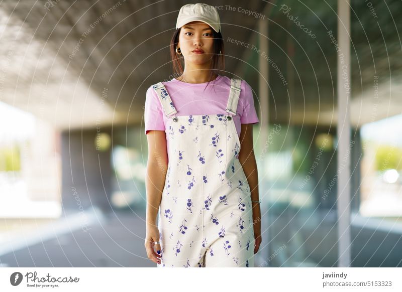 Stylish Asian woman walking towards camera style appearance modern building street urban outfit portrait female young asian ethnic t shirt overall cap