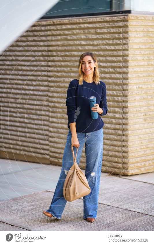 Merry female against modern building woman street style smile outfit daytime bag thermos pavement sweater jeans urban structure positive construction sidewalk