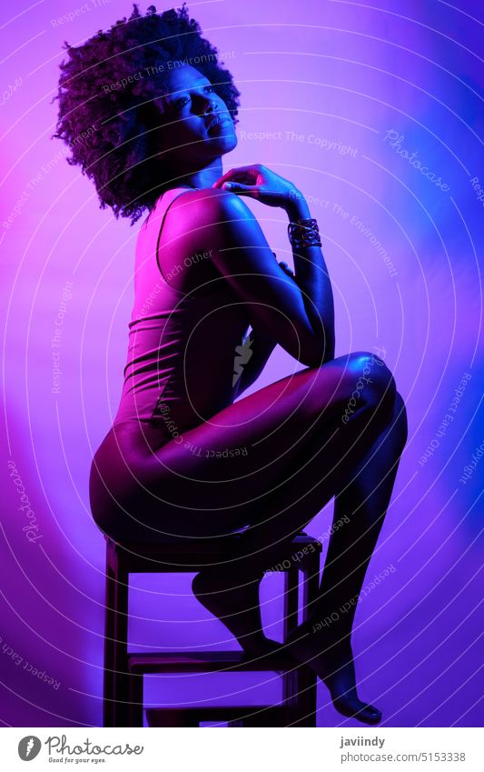 Sensual black woman under neon light touch shoulder sensual violet light stool model slim bodysuit illuminate female african american ethnic vivid sit