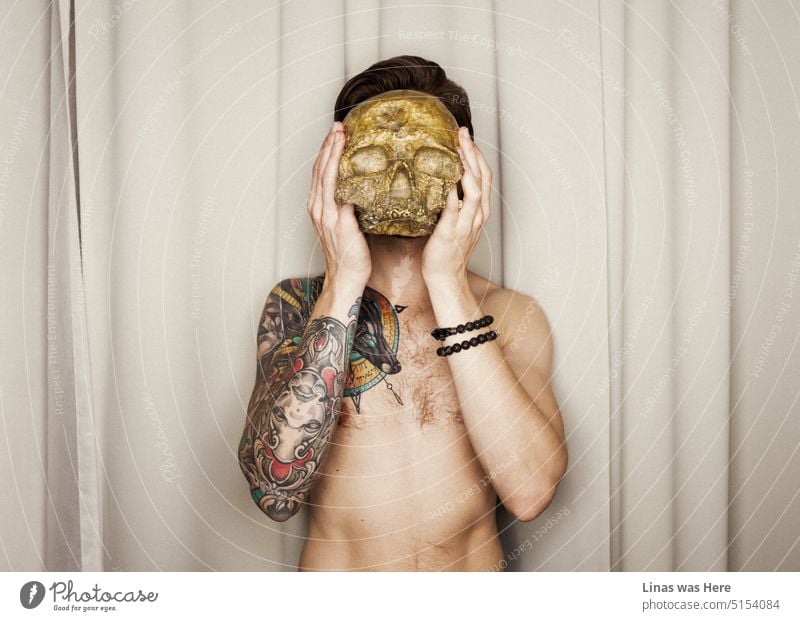 This is awkward. A male model is holding a golden skull in front of his head. His inked body, hairy chest, and some extra accessories suggest that he’s just too cool for school. Or a skull.