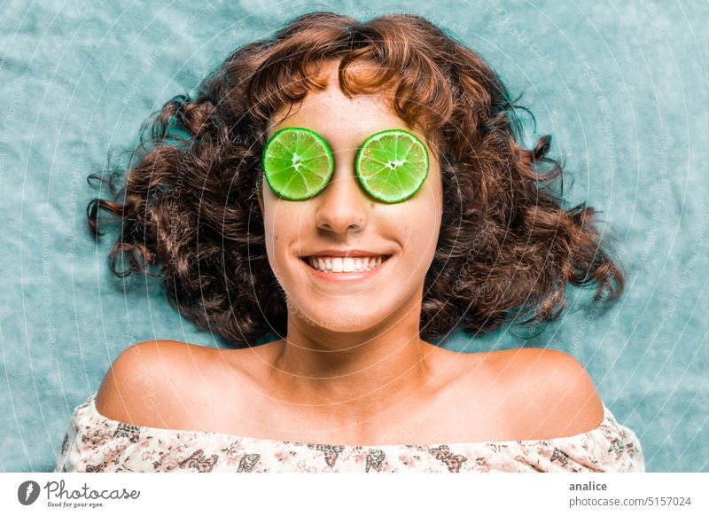 Young girl smiling with eyes covered with lime slices Girl Lemon Green Curly hair Shoulder smile teenager Teeth tan blue background Fruit citric Hair