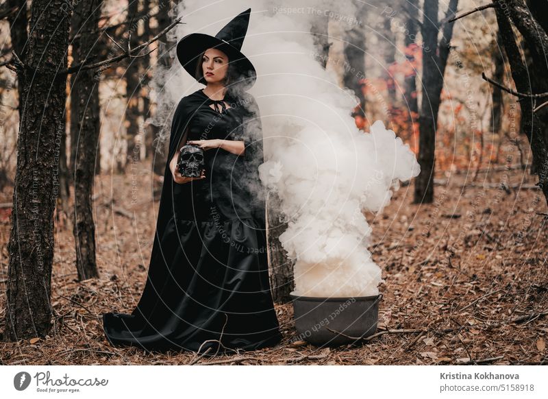 Black witch holding skull in hands, stroking it on autumn forest background. alchemy angry art bad beautiful beauty black burning carnival cast cauldron