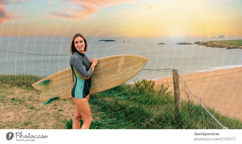 Surfer woman with wetsuit posing with surfboard looking at camera near the beach surfer young looking camera sunset sunlight smiling copy space coast