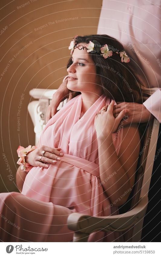 pregnant woman in a pink dress sitting in a chair behind a smiling man standing. expecting a baby together beautiful pregnancy mother love tummy happy child