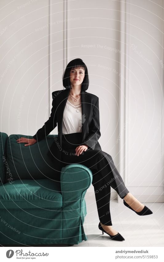 Beautiful business woman in black suit and white blouse is sitting on the green sofa at office, charming brunette female with short hair is looking in camera, girl is working indoors