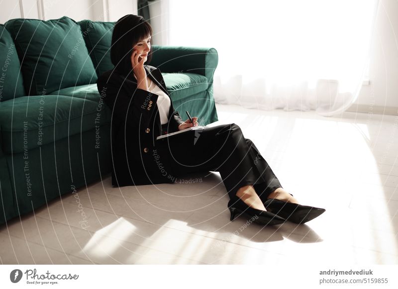 Businesswoman is talking on a smartphone. Tired brunette woman is sitting close to green sofa in a business black suit and high heels shoes with notebook. Green houseplant in interior. Work from home.