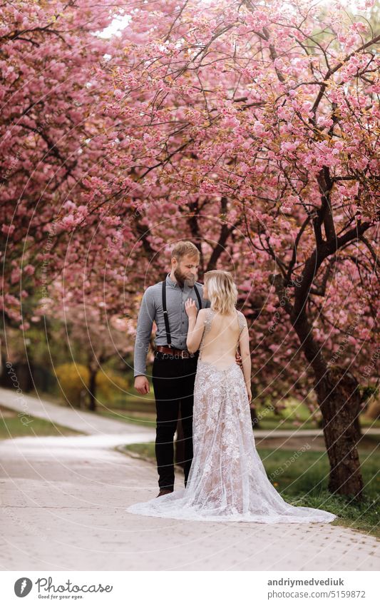 Beautiful, cheerful and lively newlyweds, groom and bride are hugging near the blooming pink cherry blossom. Wedding portrait of a close-up of a smiling bearded groom and a cute bride.
