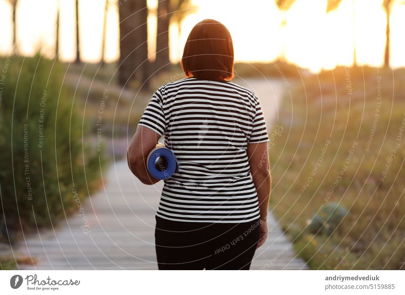 back view of active overweight woman with yoga mat walking to beach to practice yoga, exercising outdoors on sunrise in the morning. Concept of healthy lifestyle, wellbeing and fitness on nature.