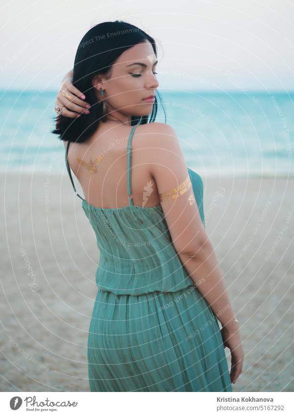 Outdoor fashion portrait of beautiful brunette lady at beach with flash tattoos on hands, back and forehead. Gypsy boho style. Woman in long green dress looking at camera.